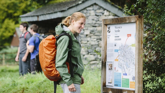 A hiker stops to look at our welcome map at Seatoller car park on a summers day while other hikers chat in the background